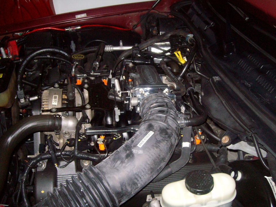 Engine bay with hoses, wires, and engine components visible. Some text on a hose