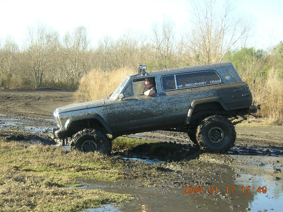 Mud-covered SUV with lifted suspension and large tires driving through a muddy field. A person is visible in the driver's seat