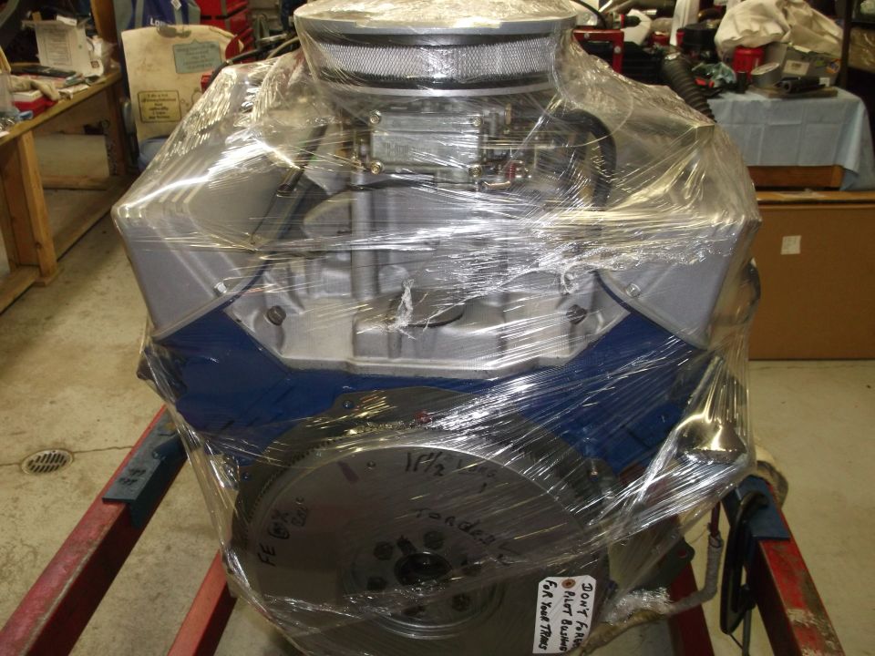 Engine block covered in plastic wrap, with a carburetor and air filter on top. Text on flywheel reads '1/2' and 'Ford'