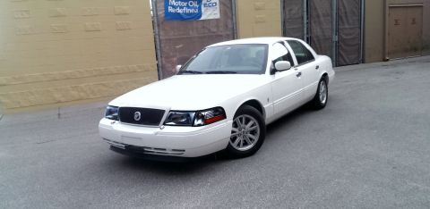 2000 Mercury Grand Marquis - A white Mercury Grand Marquis is parked on a paved surface in front of a building.