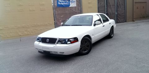 2000 Mercury Grand Marquis - A white sedan with black wheels is parked outdoors near a building.