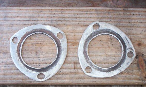 Pair of bare metal three-bolt exhaust collector flanges on weathered wood planks