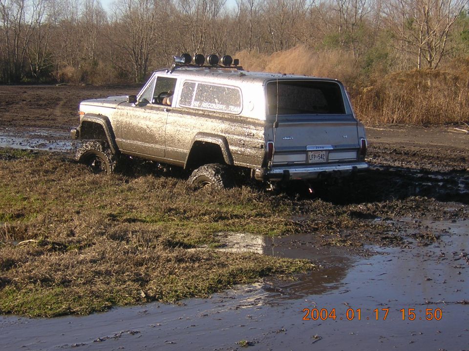 Muddy silver two-door full-size Jeep with roof light bar sitting in a mud pit