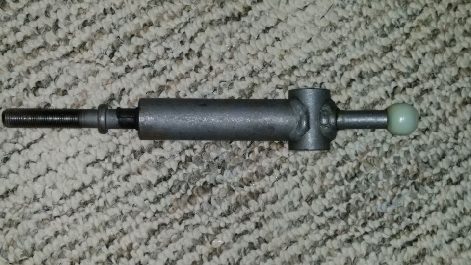 Metal gear shift linkage part with a threaded end and a light-colored ball on the other end, resting on a carpeted surface
