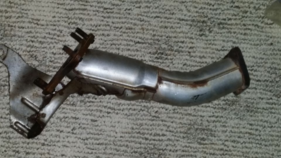 Exhaust pipe with rust and a mounting bracket, lying on a carpeted surface