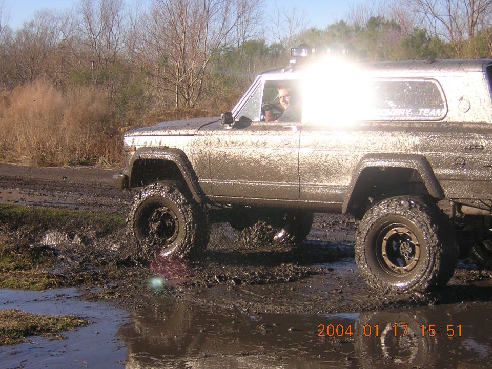 Mud-splattered SUV with off-road tires stuck in mud, driver visible, text on rear window
