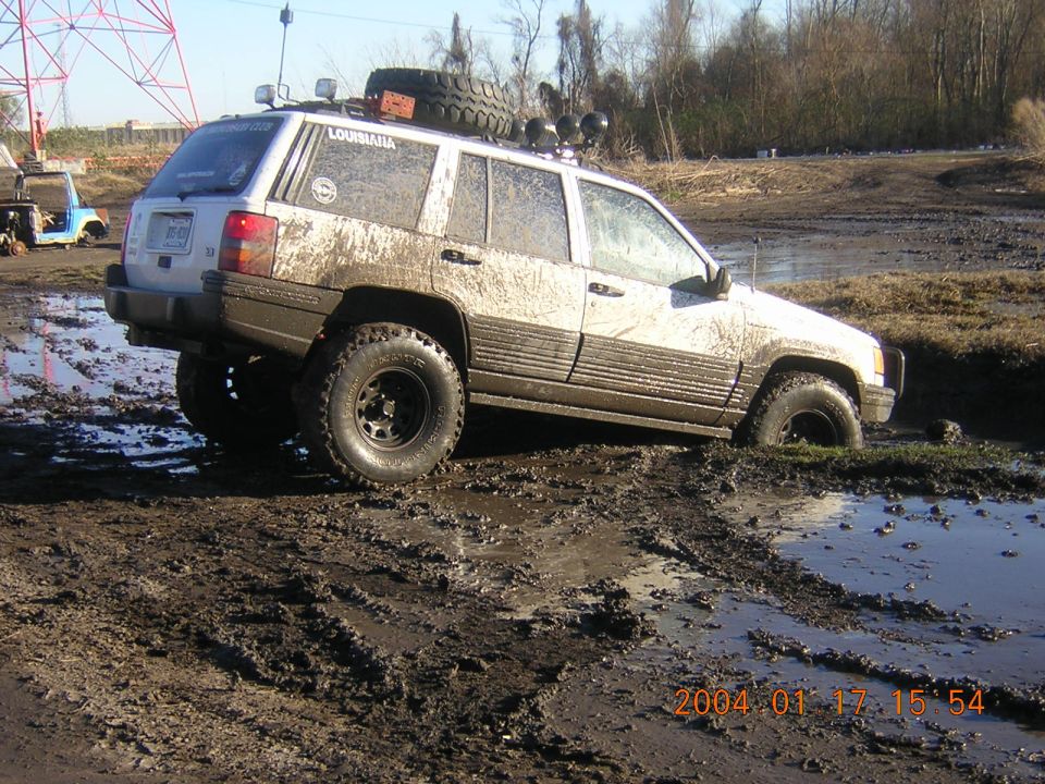 Mud-covered white Jeep Grand Cherokee with off-road tires stuck in a muddy puddle, spare tire on roof