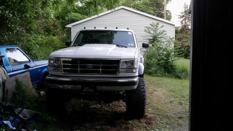 1995 Ford F-350 - A lifted white Ford pickup truck is parked in front of a garage, with another blue truck partially visible to its left.