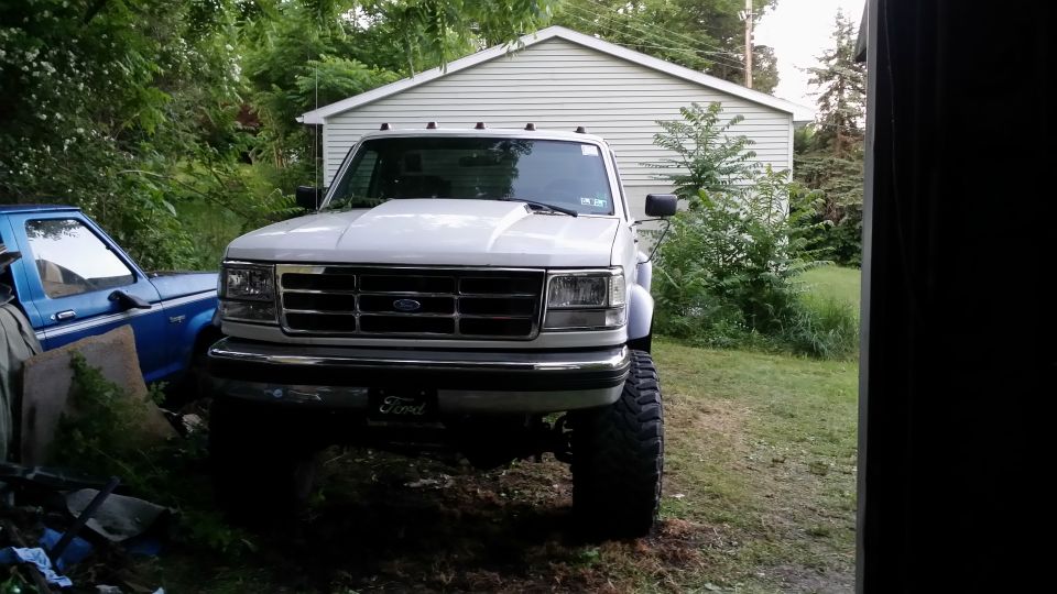 Lifted white Ford pickup truck parked in front of a garage, with a blue truck to the left. The Ford appears to be a 1995 model