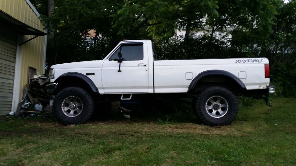 White pickup truck with lifted suspension and aftermarket wheels, parked on grass next to a building