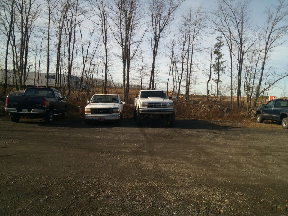 White pickup truck with a lifted suspension parked in front of bare trees. Another white pickup and a black pickup are also visible