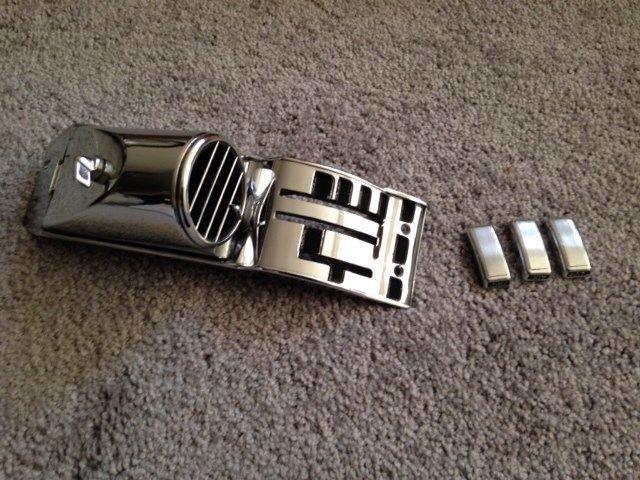 Chrome vent trim piece with horizontal slats and three small chrome emblems on a carpeted surface