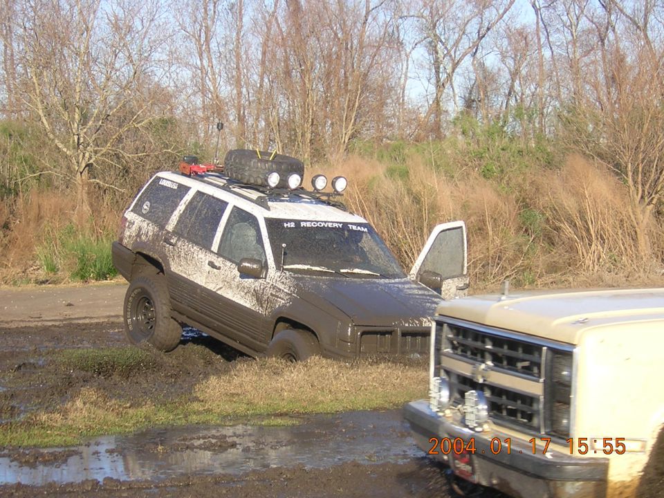 Muddy SUV stuck in a ditch with its door open, another vehicle's front end visible on the right