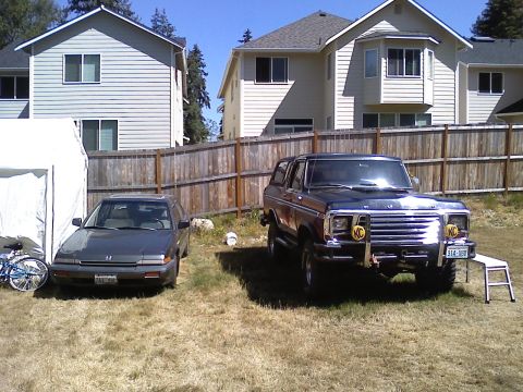 1979 Ford Bronco - A dark blue Ford Bronco and a gray sedan are parked in a grassy yard with houses in the background.