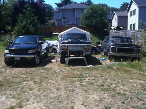 1979 Ford Bronco - Three Ford trucks are parked in a yard. The central vehicle, a 1978 Ford Bronco, is elevated on jack stands.