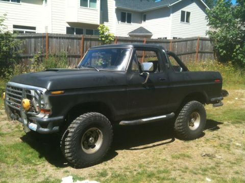 1979 Ford Bronco - Flat-black full-size Ford Bronco with brush guard, winch and oversized mud tires parked on grass by a fence.