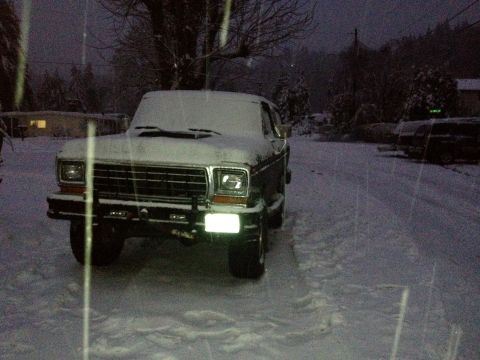 1979 Ford Bronco - A Ford Bronco sits in the snow at dusk, its headlights illuminating the snowy road.