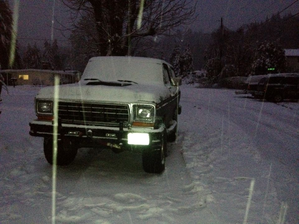 Dark green Ford Bronco covered in snow, front view. Headlights are on