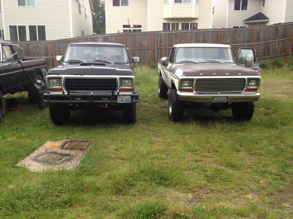 Two Ford trucks parked in a grassy yard. The one on the right is a 1979 Ford Bronco