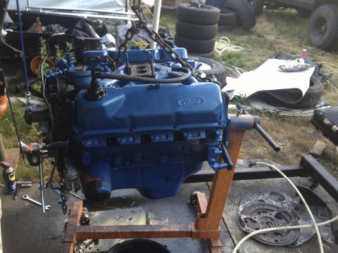 1979 Ford Bronco - A blue Ford V8 engine is suspended from chains on an engine stand, with various parts and tires in the background.