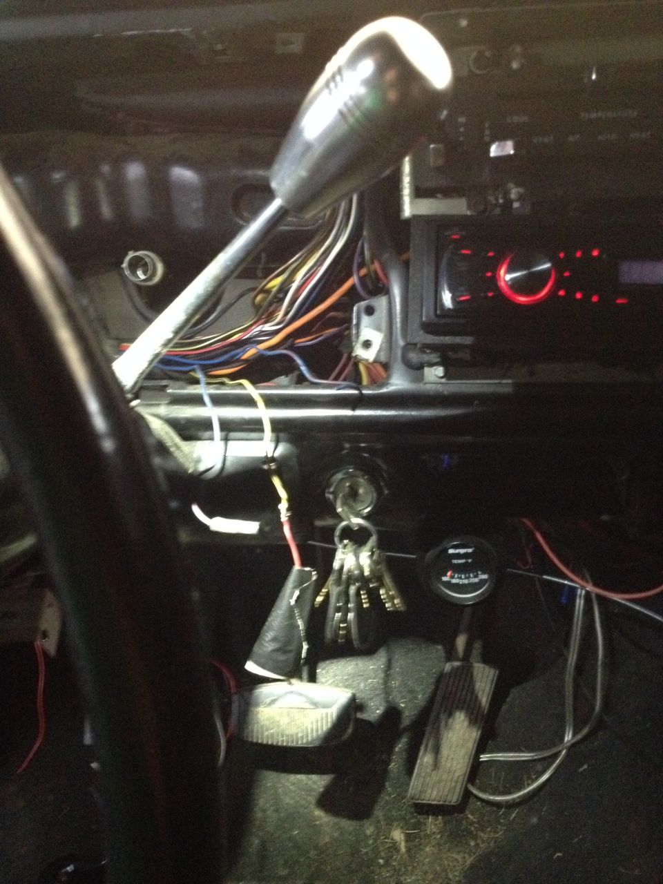 Car interior showing dashboard, steering column, gear shifter, exposed wiring, and pedals. Aftermarket radio installed