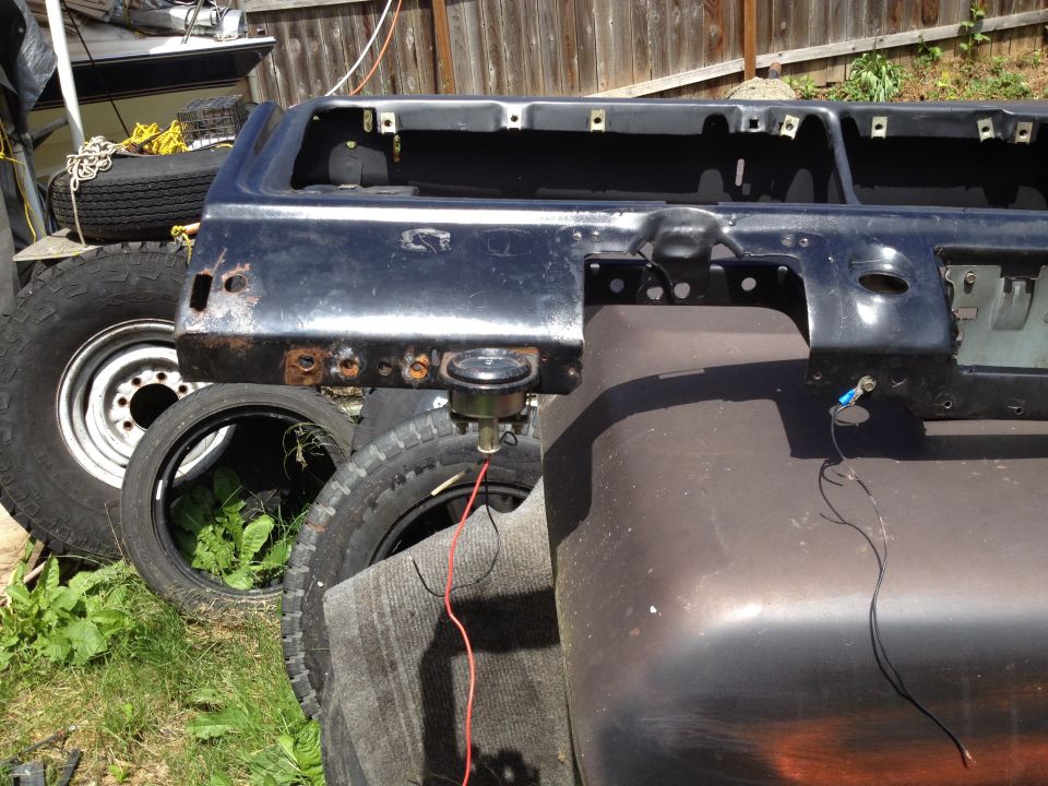 Black vehicle body panel with rust spots and exposed metal, resting on tires. A boat is visible in the background