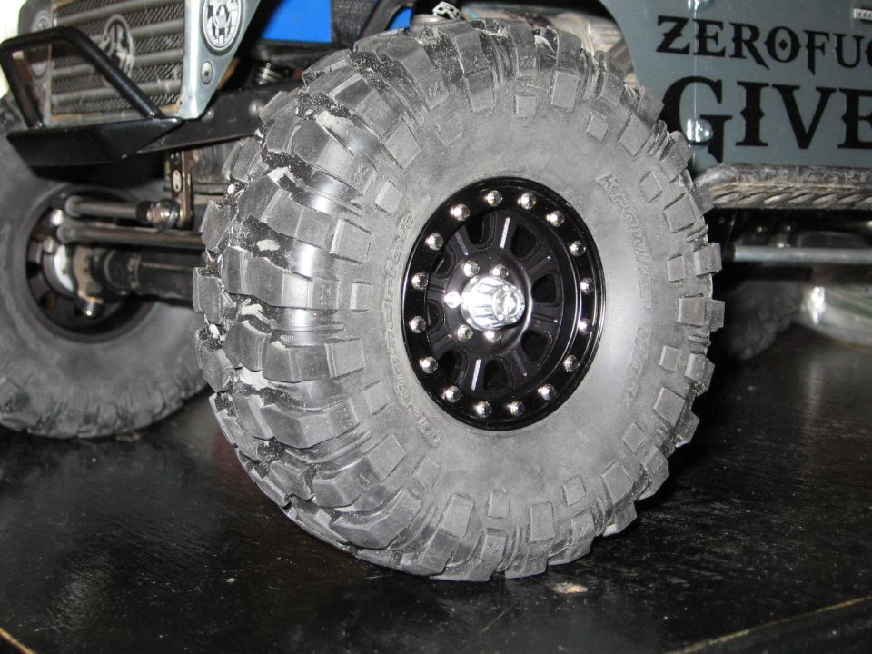 Close-up of a large, knobby RC car tire on a black beadlock rim. The vehicle has a winch and a grille visible
