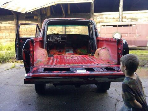 1978 Ford Bronco - A young boy looks at the open bed and interior of a red pickup truck, which appears to be in a state of disrepair.