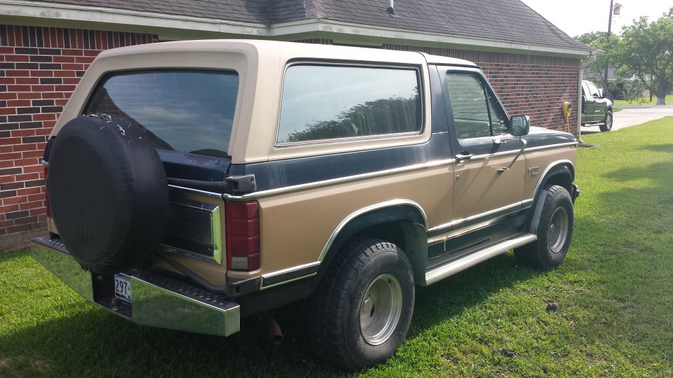 Black and tan two-tone Ford Bronco SUV with rear spare tire cover, parked on grass by a brick house