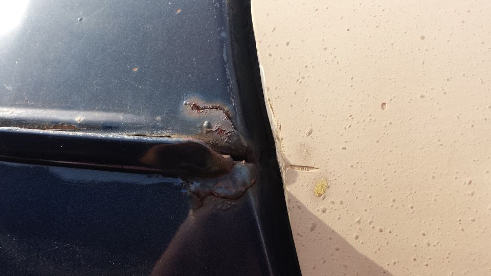 Close-up of rust bubbles and corrosion on dark blue Bronco body panel seam beside tan panel