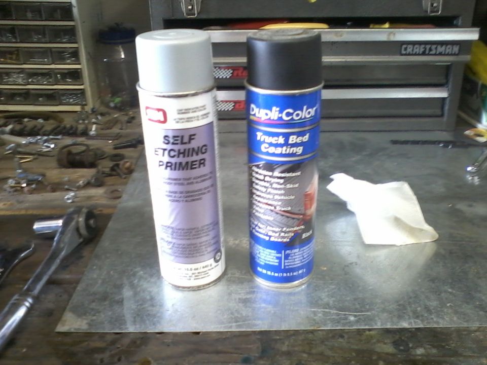 Two spray cans, one labeled 'SELF ETCHING PRIMER' and the other 'Dupli-Color Truck Bed Coating', sit on a metal surface in a workshop