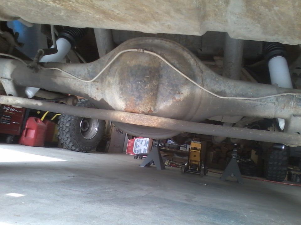 Underside view of a vehicle's rear axle, differential, shocks, and brake lines. Tires and shop equipment are visible in the background