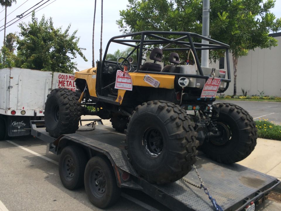 Yellow off-road vehicle with large tires on a trailer, has a roll cage and two seats. 'For Sale' signs are visible