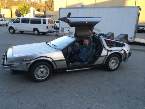 1968 Ford Mustang - A man sits in a DeLorean DMC-12 with its iconic gull-wing doors open, parked outdoors.