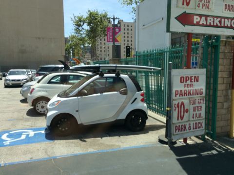 1968 Ford Mustang - A white Smart car with a surfboard strapped to its roof rack is parked next to other vehicles.