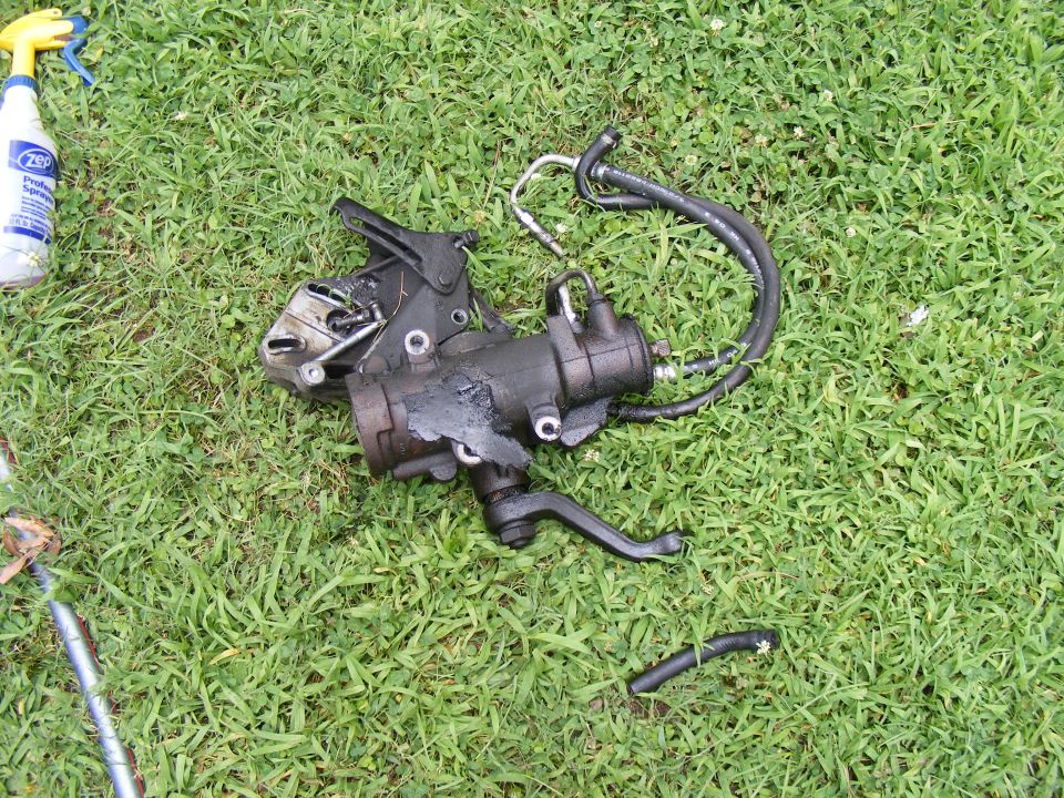 Black steering gear box with hoses lying on grass, next to a spray bottle