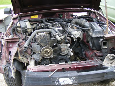 Engine bay of a damaged maroon vehicle, showing engine components and front-end damage. Tools are on the frame
