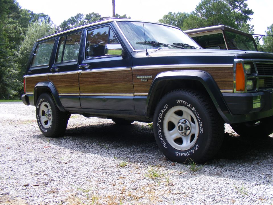 Jeep Grand Wagoneer with wood paneling parked on gravel, another vehicle partially visible behind it