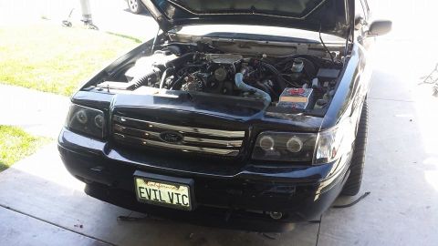 2003 Ford Crown Victoria - Black Ford sedan with hood up showing the chrome grille and a California 'EVIL VIC' front license plate.