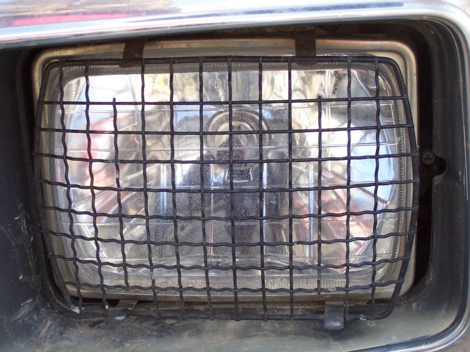 Rectangular headlight with black wire mesh guard in chrome bezel, covered in dust