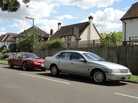 1999 Ford Fairlane - Two cars, a silver sedan and a red Ford Mustang, are parked on the side of a street in front of houses.