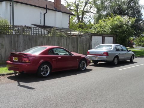 1999 Ford Fairlane - Two cars are parked on the side of a street in front of a house and garages.