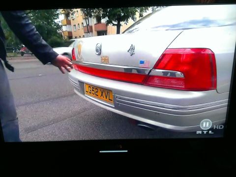 1999 Ford Fairlane - A person's hand reaches towards the rear of a silver sedan, which displays various decals and a UK license plate.