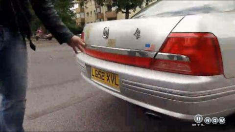 1999 Ford Fairlane - A person's hand reaches towards the rear of a silver sedan, revealing its taillights and license plate.