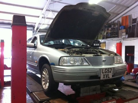 1999 Ford Fairlane - A silver sedan is raised on a car lift with its hood open, revealing the engine bay.