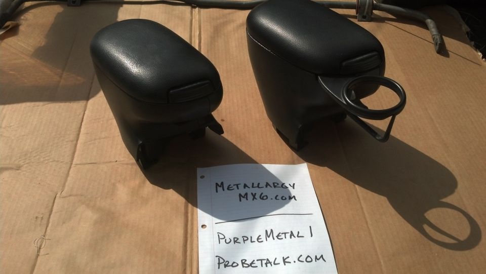 Two black car armrests with integrated cup holders, one with a ring holder. Paper with text 'METALLARGY MX6.com', 'PurpleMetal I', 'PROBETALK.COM' is nearby