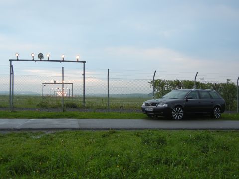 2003 Audi A6 - A dark Audi A6 Avant wagon sits parked next to a chain-link fence with airport runway lights in the background.