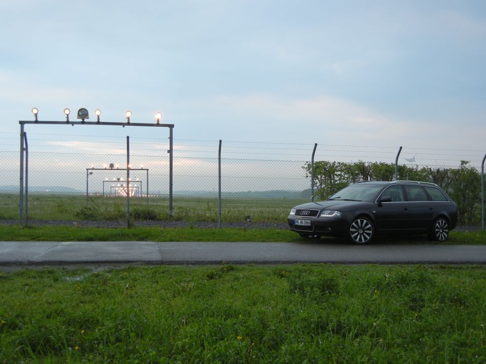 Dark Audi A6 Avant parked by a fence near an airport runway. License plate is visible