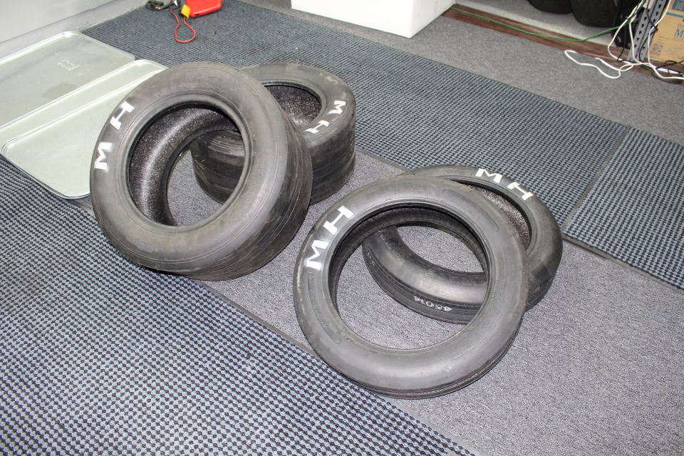 Four M&H drag racing slick tires arranged on a gray carpeted floor