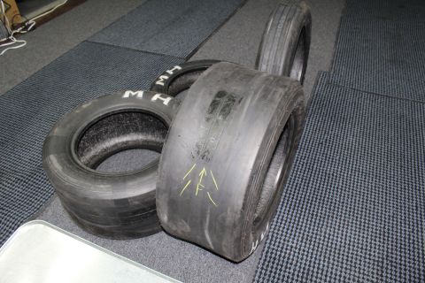 2003 Ford Crown Victoria - wide bald drag slick with yellow arrow marks rests beside narrower "M H" lettered tires on a gray floor.